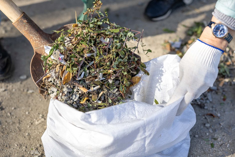 A gardener wearing white gloves and a wristwatch is collecting garden debris, including small branches, leaves, and gravel, with a trowel into a large white garden waste bag. The scene takes place out